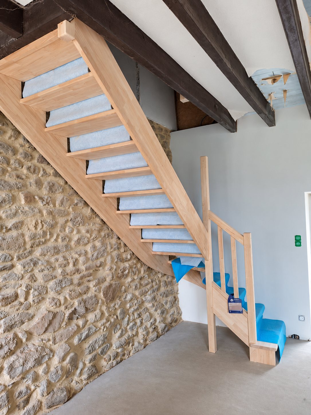 Un escalier en bois clair avec marches protégées par un film bleu et gris, installé le long d'un mur en pierres apparentes dans une maison en rénovation.