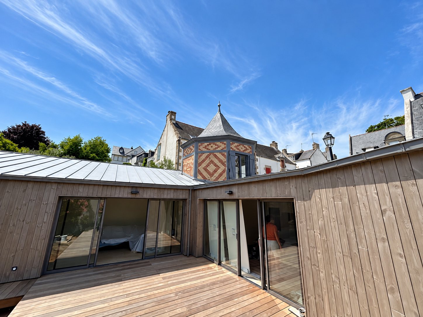 Extension en bardage bois et zinc avec terrasse, devant la tourelle en briques.