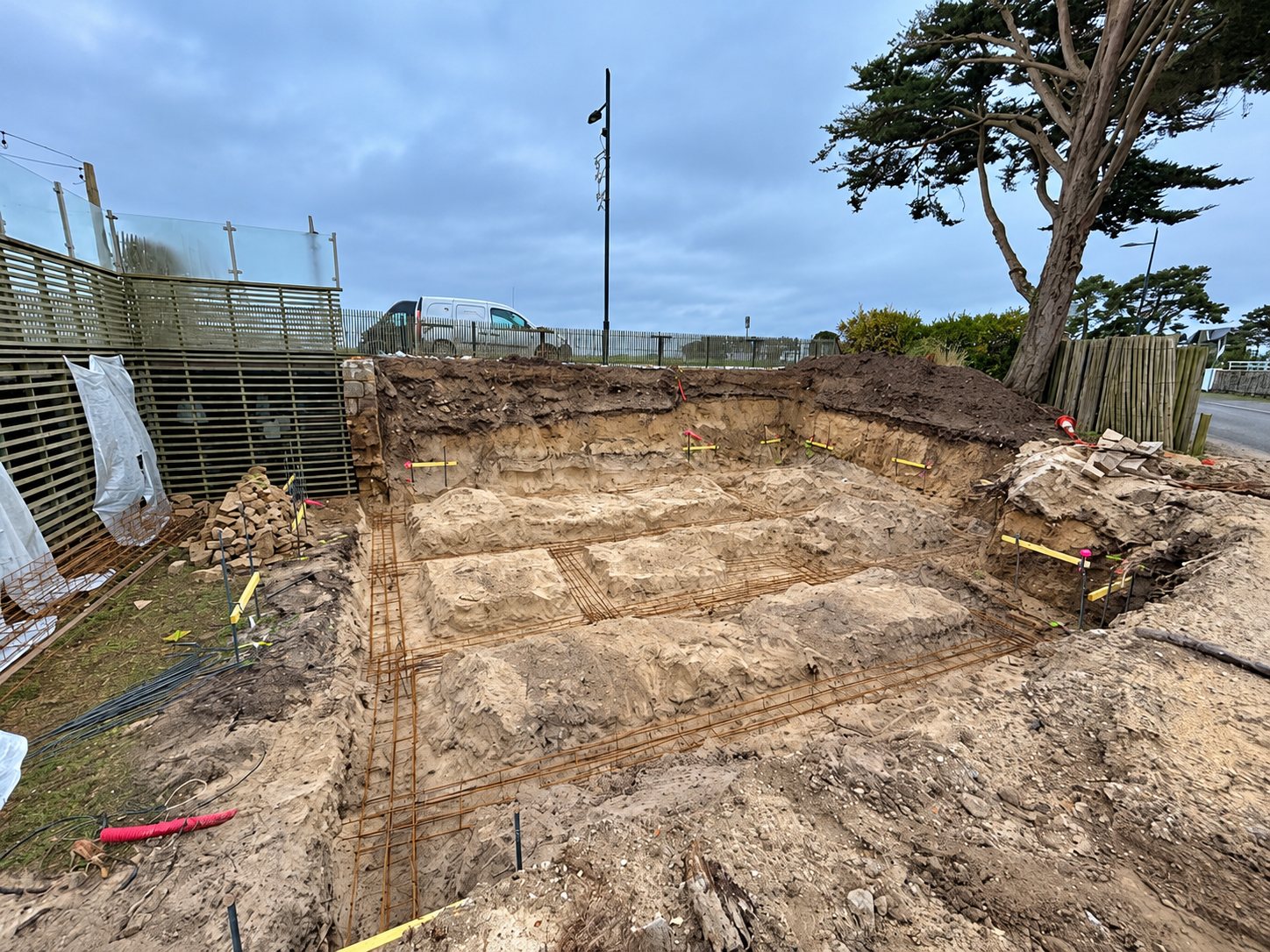 Chantier de terrassement avec une fouille sableuse et des armatures métalliques posées au fond, près d'une clôture, d'un cyprès et d'une camionnette blanche.