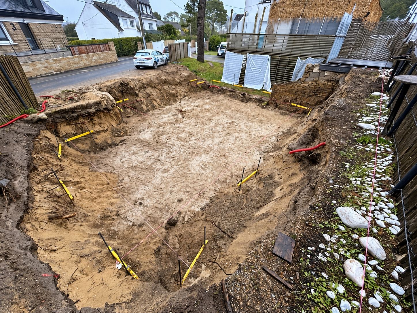 Grande excavation rectangulaire dans un sol sableux, délimitée par des piquets jaunes et des gaines rouges, devant une rue résidentielle bordée de maisons.