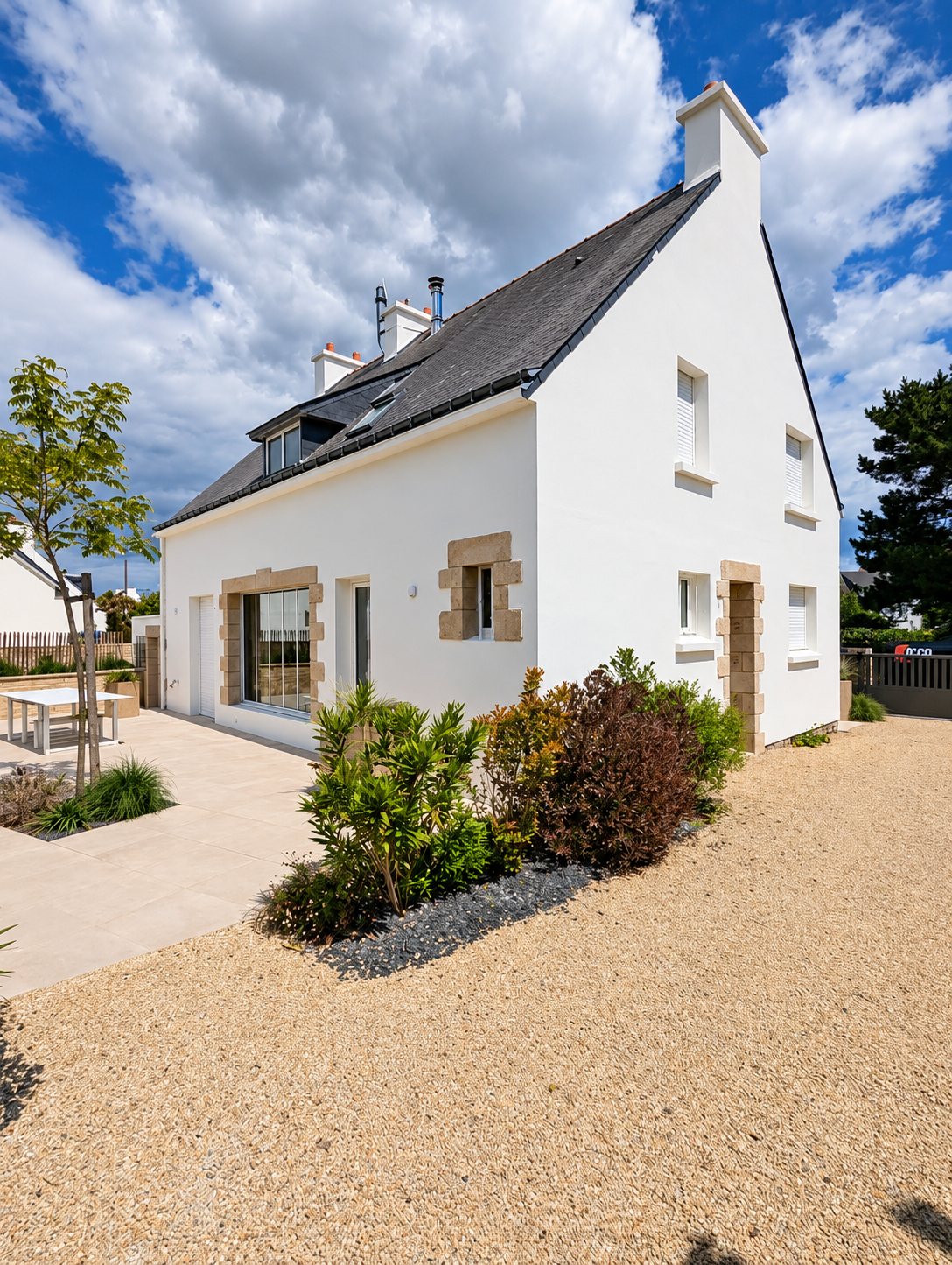 Maison rénovée aux murs blancs, toit d'ardoise et encadrements de fenêtres en pierre, entourée d'un jardin avec gravier et terrasse claire.