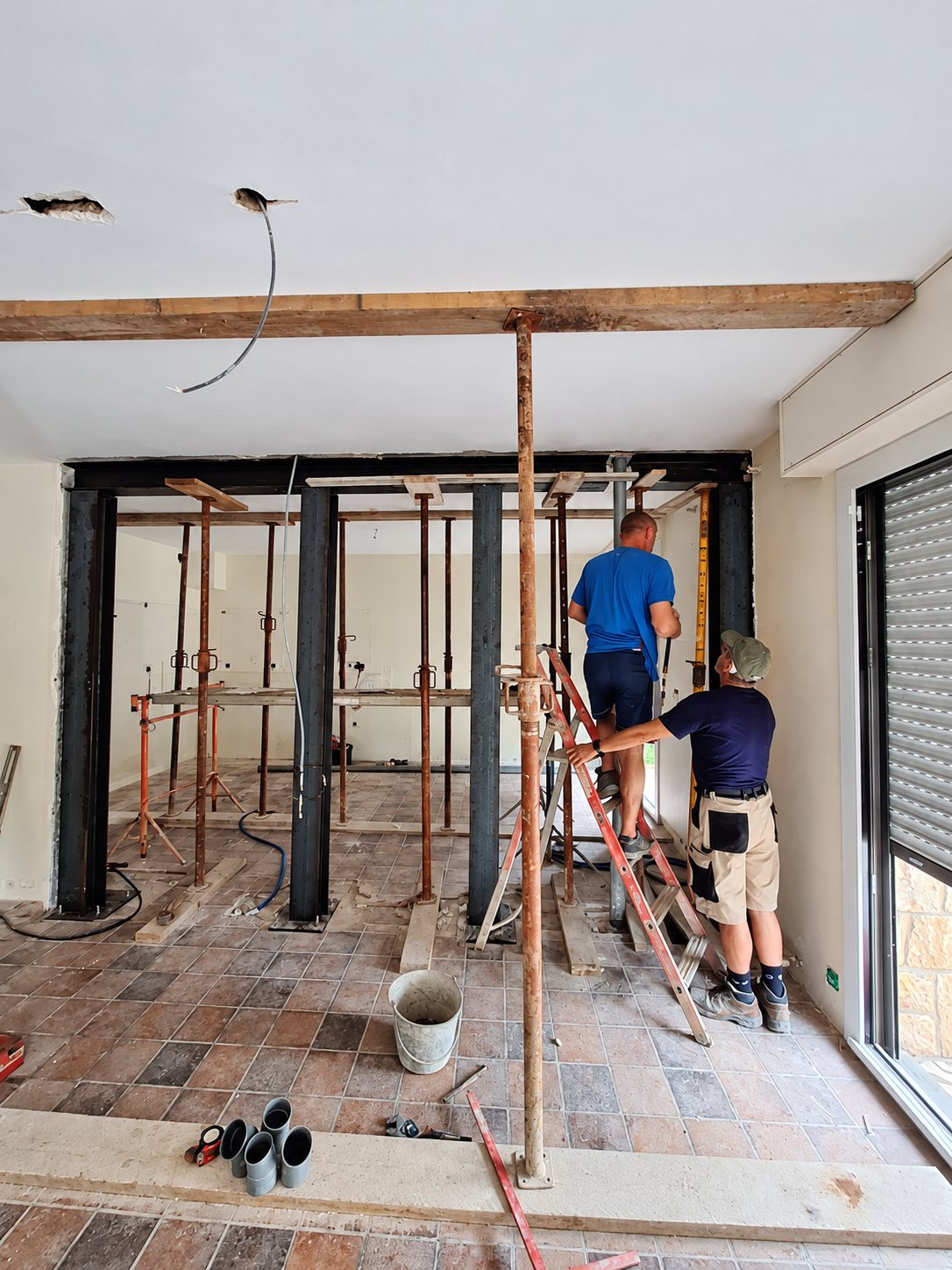 Deux ouvriers travaillent dans une pièce en rénovation, soutenue par des étais et des poteaux en acier, l'un d'eux monté sur une échelle.