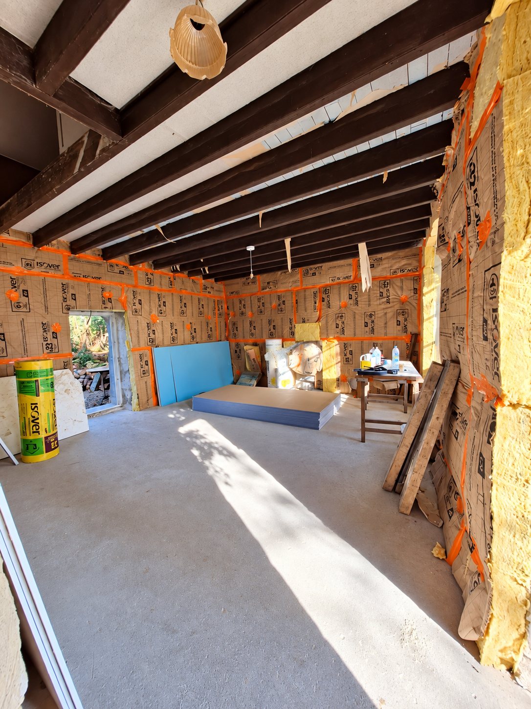 Intérieur d'une pièce en rénovation avec murs isolés par des panneaux Isover, poutres sombres au plafond et plaques de plâtre empilées au sol.