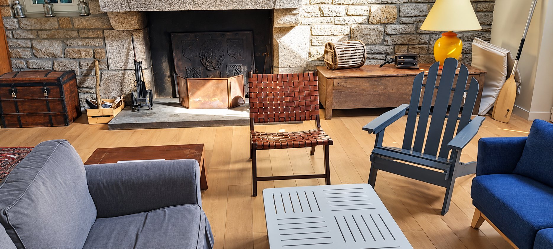 Un salon lumineux avec une grande cheminée en granit, un fauteuil en cuir tressé, un fauteuil Adirondack bleu-gris, des canapés et une table basse sur un parquet clair.